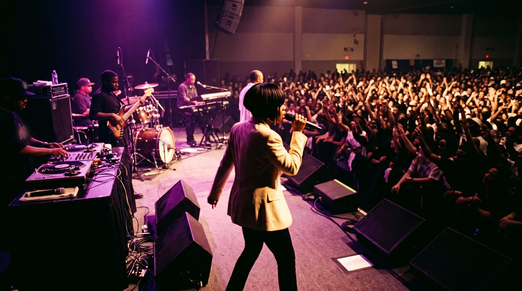 The singer in a cream blazer, seen from behind on stage, faces a large crowd with raised hands while the band plays behind her